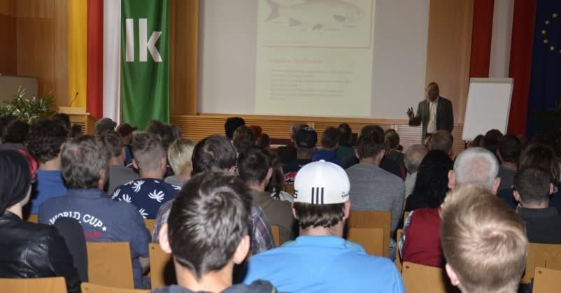 Ein Mann hält eine Präsentation für ein Publikum in einem Konferenzsaal. Er steht vor einer Leinwand mit Text und einer Tafel. Viele Menschen sitzen in Reihen vor dem Redner.