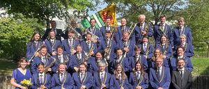 Ein großes Orchester posiert mit Instrumenten wie Trompeten, Saxophonen und Flöten für ein Foto. Sie tragen blaue Uniformen mit Krawatten und stehen auf einem Kopfsteinpflaster. Bäume und eine Flagge sind im Hintergrund.