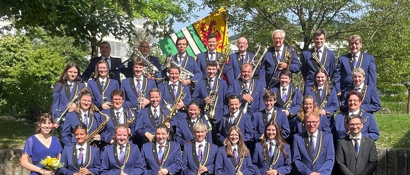Ein großes Orchester posiert mit Instrumenten wie Trompeten, Saxophonen und Flöten für ein Foto. Sie tragen blaue Uniformen mit Krawatten und stehen auf einem Kopfsteinpflaster. Bäume und eine Flagge sind im Hintergrund.