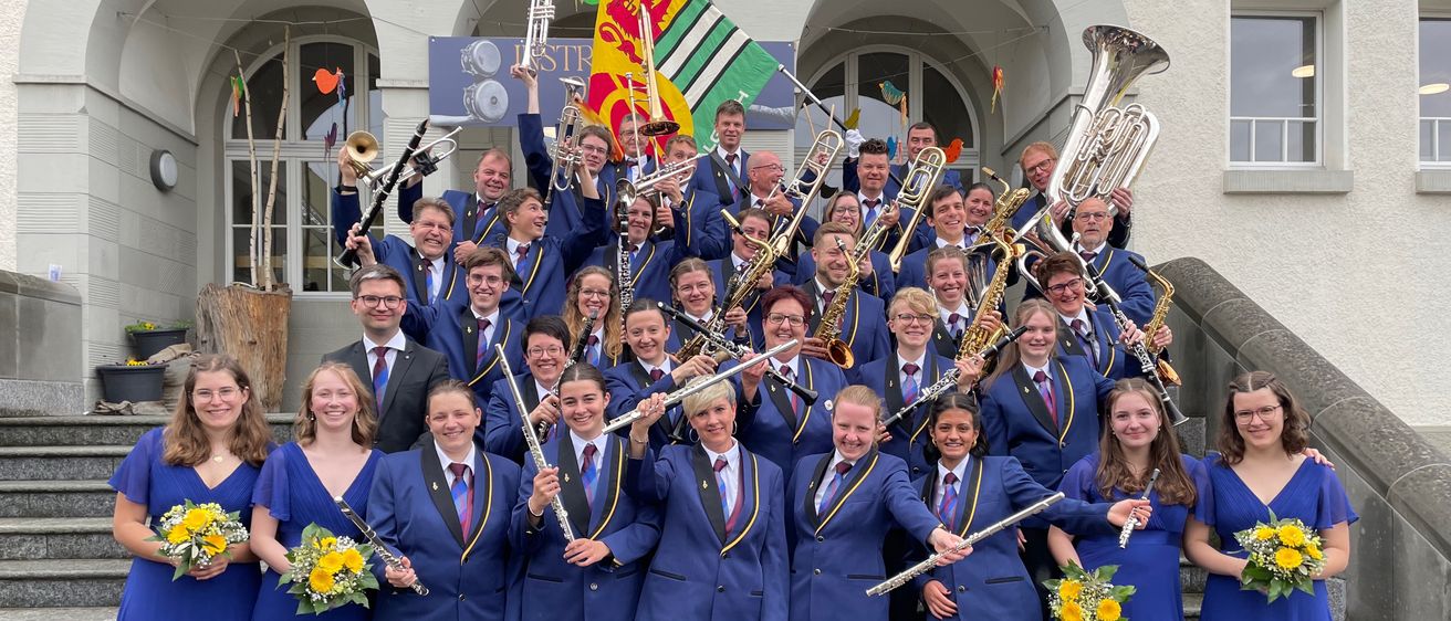 A band of musicians dressed in blue uniforms stands on the steps of a building, holding various musical instruments, including trumpets, saxophones, and tubas, with a flag in the background.