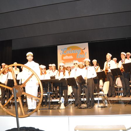 Eine Gruppe von Menschen in Matrosenuniformen steht auf der Bühne, einige spielen Instrumente. Ein großes Steuerrad steht davor. Ein Banner mit den Worten 'Leinen los!' im Hintergrund.