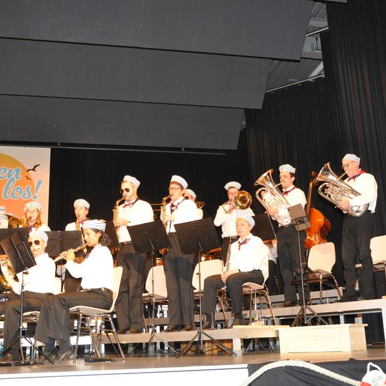 A band is performing on stage, wearing sailor hats and uniforms. Some sit on chairs while others stand, playing various instruments. The stage has a black backdrop and chairs.