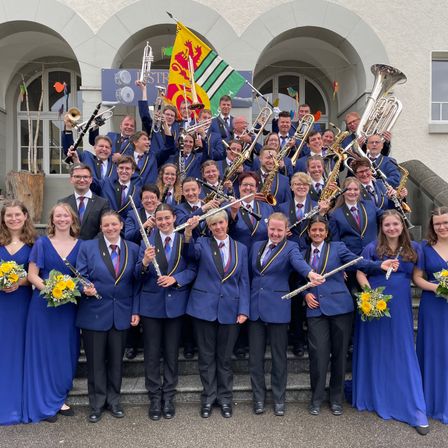 Ein Musikorchester mit verschiedenen Instrumenten, darunter Trompeten, Posaunen und Flöten, posiert für ein Foto vor einem Gebäude. Sie tragen blaue Uniformen und halten Blumen.