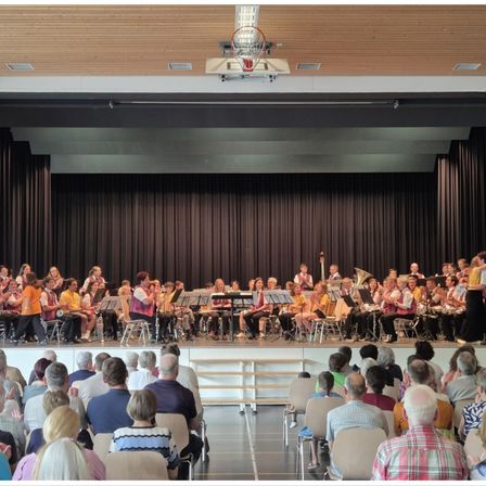 Ein großes Orchester spielt auf der Bühne mit verschiedenen Instrumenten, während ein Publikum in Stühlen davor sitzt.