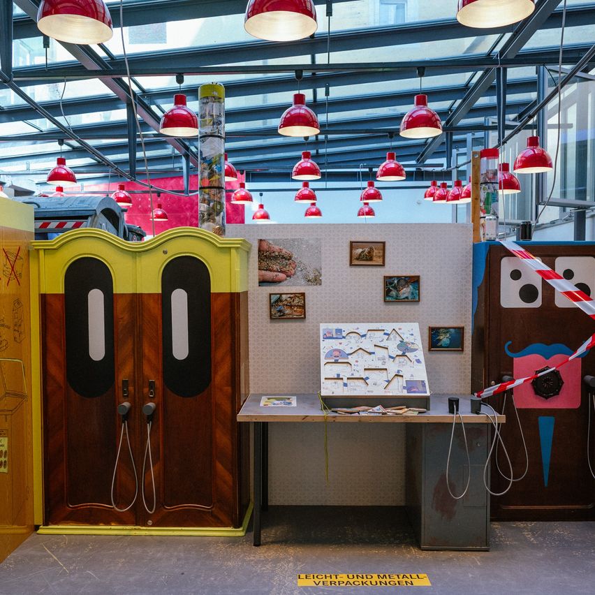 Ein Raum mit einem gelben und braunen Schrank mit großen Augen. Rote Lampen hängen von der Decke. Ein Tisch hat ein Spielbrett darauf. Ein gelbes Schild weist auf leichte und Metallverpackungen hin.