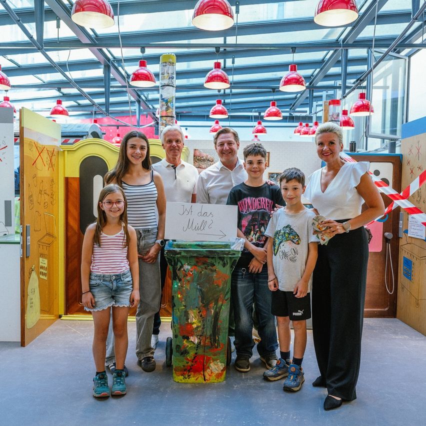 Eine Familie mit sieben Personen steht in einer bunten Umgebung mit einem Schild, auf dem 'Abfall in Mühle 2' steht. Die Familie besteht aus zwei Frauen, vier Jungen und einem Mann. Sie lächeln für ein Foto.
