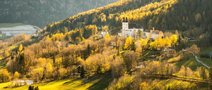 Ein Blick vom Hügel auf ein Dorf mit einer großen Kirche, umgeben von herbstlichen Bäumen und einer Kulisse aus Bergen unter einem hellen Himmel.