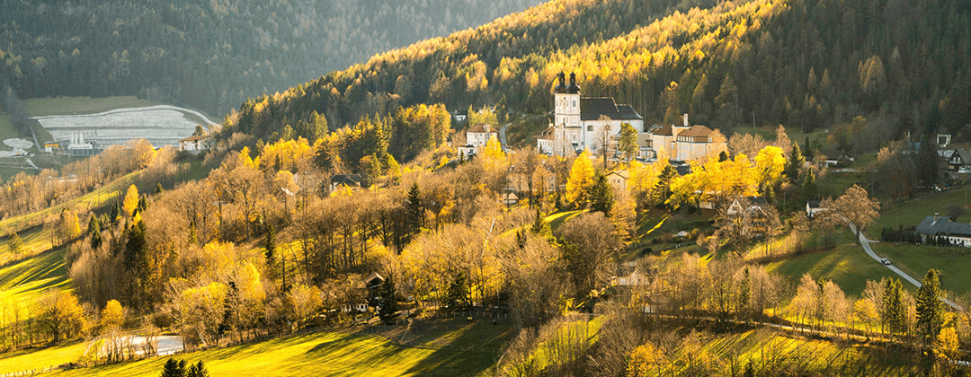 Ein Blick vom Hügel auf ein Dorf mit einer großen Kirche, umgeben von herbstlichen Bäumen und einer Kulisse aus Bergen unter einem hellen Himmel.