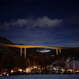 Ein Nachtblick auf eine Brücke über einem Tal mit Schnee und Bäumen, beleuchtet von Lichtern.