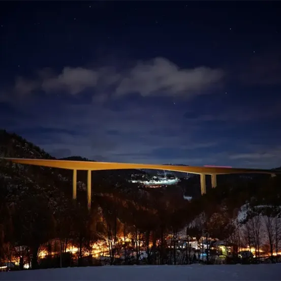 Ein Nachtblick auf eine Brücke über einem Tal mit Schnee und Bäumen, beleuchtet von Lichtern.