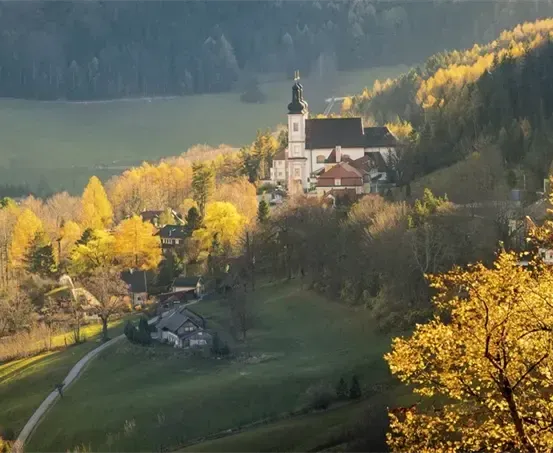Eine große Kirche mit Turm steht auf einem Hügel, umgeben von herbstlichen Bäumen. In der Nähe sind Häuser und ein kleiner See.