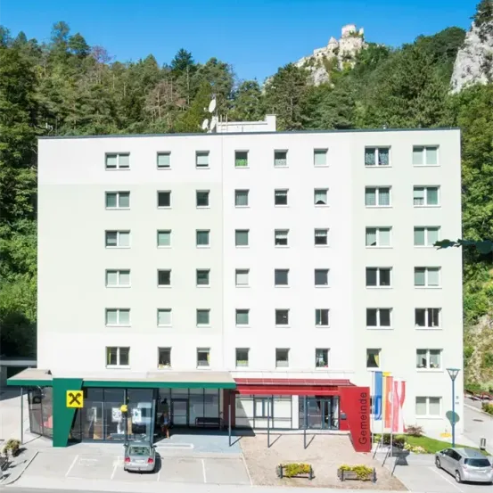Ein weißes Gebäude mit mehreren Fenstern steht vor einem Berg und ein Auto ist davor geparkt. Das Gebäude hat ein rotes Vordach und ein grünes und weißes Vordach.