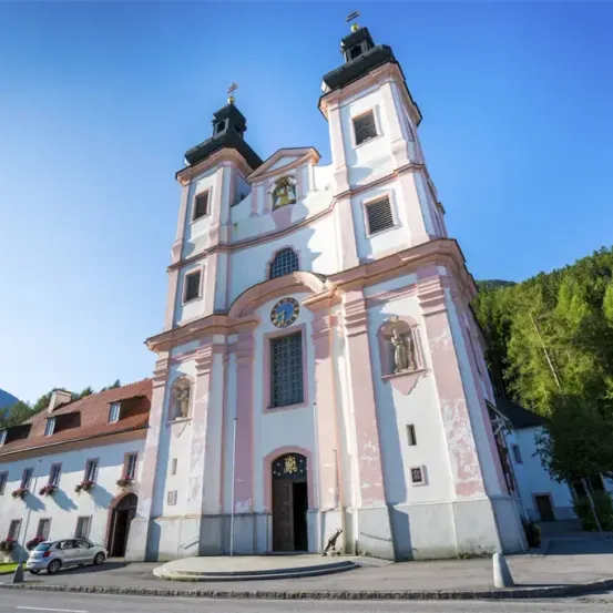 Eine Kirche mit zwei Türmen, einer Uhr und einer Statue, umgeben von Bäumen und Bergen. Ein Auto ist davor geparkt.