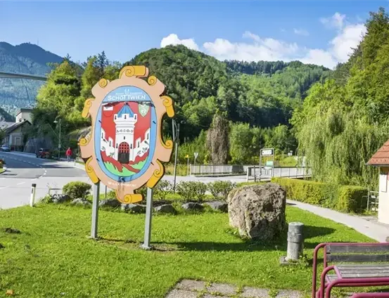 Ein Schild mit einem Burglogo steht in einem Grasbereich, in der Nähe eines Hauses und einer Brücke, mit Bergen in der Ferne.