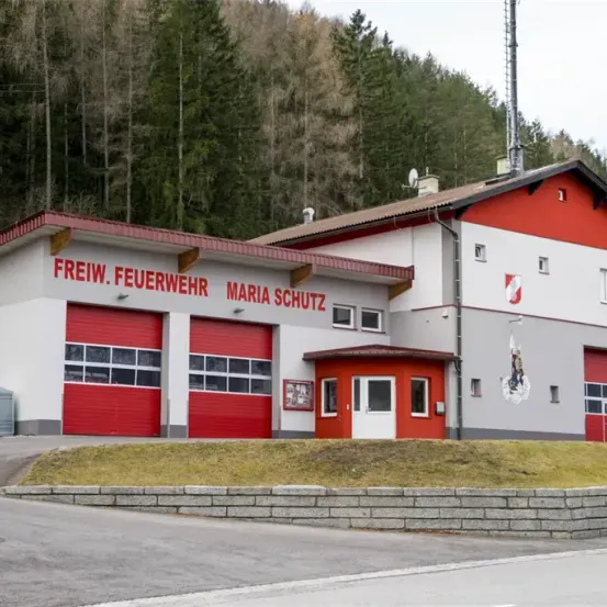 Ein rotes und weißes Gebäude mit der Aufschrift Freiw. Feuerwehr Maria Schutz mit zwei roten Garagen und einer Flagge.