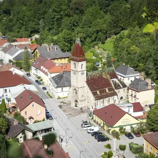 Ein Luftbild eines Dorfes mit vielen Häusern und einer Kirche mit Turm in der Mitte. Autos sind auf der Straße vor der Kirche geparkt.