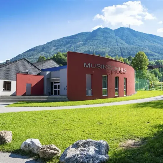 Ein rotes Gebäude mit weißen Akzenten und dem Wort Musikschule an der Seite. Es befindet sich in einer grünen Landschaft mit Bergen im Hintergrund.