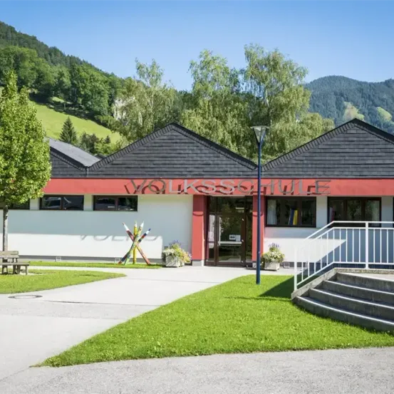 Das Volksschule-Gebäude mit rotem Rand, schwarzem Dach und Eingangsbereich ist von Bäumen und Rasen umgeben. Das Gebäude hat eine Bank links und eine Laterne rechts.