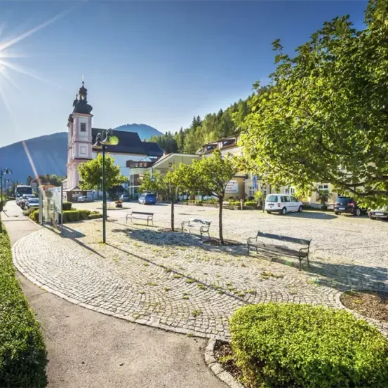 Ein Stadtplatz mit einer Kirche im Hintergrund, umgeben von Bänken, geparkten Autos und Bäumen. Die Sonne scheint hell.