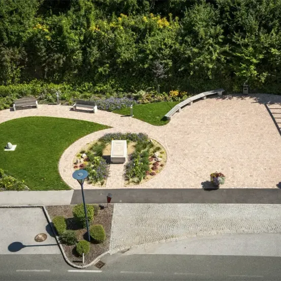 Ein Luftbild eines Gartens mit zwei hölzernen Bänken und einem zentralen Betonweg, umgeben von Grün und Bäumen. Der Garten wird von einer gepflasterten Straße begrenzt.