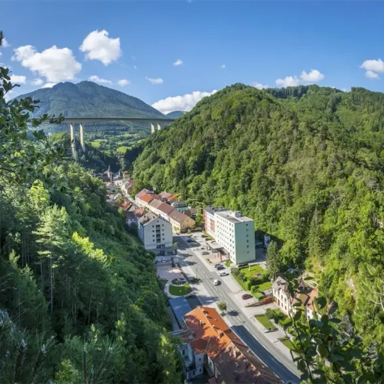 Luftaufnahme eines Dorfes zwischen Bergen mit einer Brücke, Gebäuden und einer Straße mit Autos. Viel Grün, Bäume und ein blauer Himmel mit Wolken.