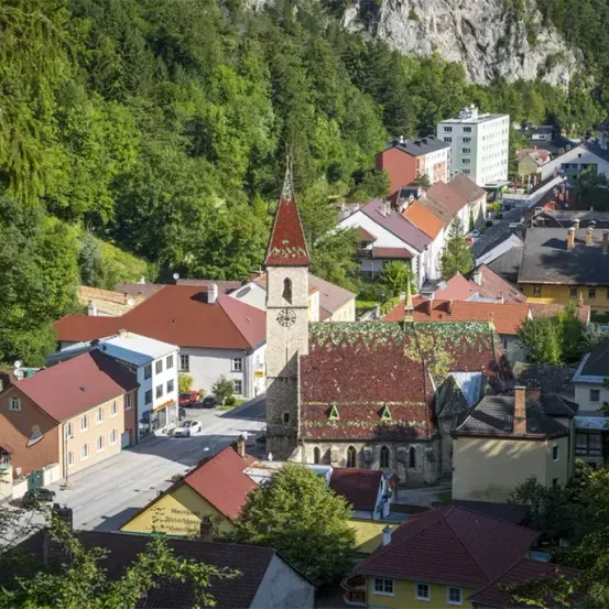 Ein malerisches Dorf in einem Tal, umgeben von Bäumen und Bergen. Mehrere Gebäude, darunter ein Turm und eine Kirche, sind sichtbar. Ein Auto ist auf der Straße geparkt, und eine Felsformation befindet sich rechts.