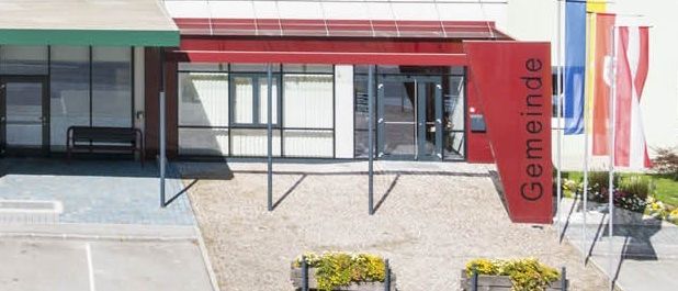 Ein weißes Gebäude mit Glasfenstern, einem roten Vordach und Fahnen draußen. Es gibt Pflanzen in Pflanzkübeln und eine Bank in der Nähe des Eingangs.