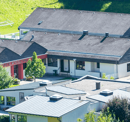 Ein Luftbild einer Schule mit einem großen Gebäude und mehreren kleineren Gebäuden. Das Gebiet ist von Bäumen und Gras umgeben. Die Schule hat ein graues Dach und rote Wände.