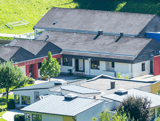 Ein Luftbild einer Schule mit einem großen Gebäude und mehreren kleineren Gebäuden. Das Gebiet ist von Bäumen und Gras umgeben. Die Schule hat ein graues Dach und rote Wände.