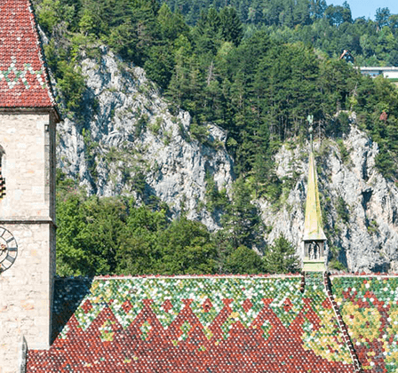 Ein Steinturm mit einer Uhr und einem gefliesten Dach steht vor einem Hintergrund aus felsiger Klippe und üppiger Grünfläche.