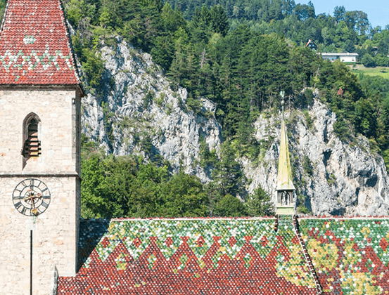 Ein Steinturm mit einer Uhr und einem gefliesten Dach steht vor einem Hintergrund aus felsiger Klippe und üppiger Grünfläche.