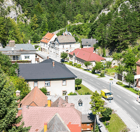Luftaufnahme einer Stadt in den Bergen mit mehreren Häusern, einige mit roten Dächern. Eine Straße verläuft durch die Stadt mit einem gelben Van und einem schwarzen Auto, die an der Seite geparkt sind.