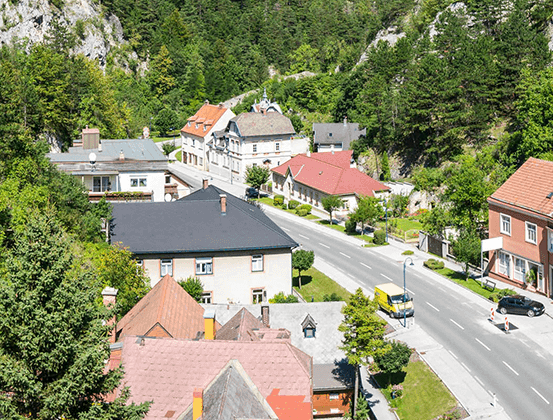 Luftaufnahme einer Stadt in den Bergen mit mehreren Häusern, einige mit roten Dächern. Eine Straße verläuft durch die Stadt mit einem gelben Van und einem schwarzen Auto, die an der Seite geparkt sind.