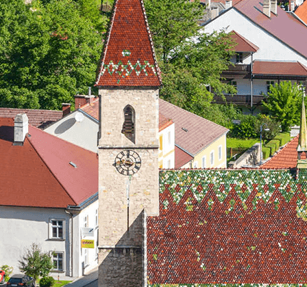 Luftaufnahme einer Stadt mit traditioneller Architektur. Rot gedeckte Dächer, ein Turm mit einer Uhr und üppige Grünflächen. Mehrere Häuser mit Schornsteinen, einige geparkte Autos und eine steile Straße.