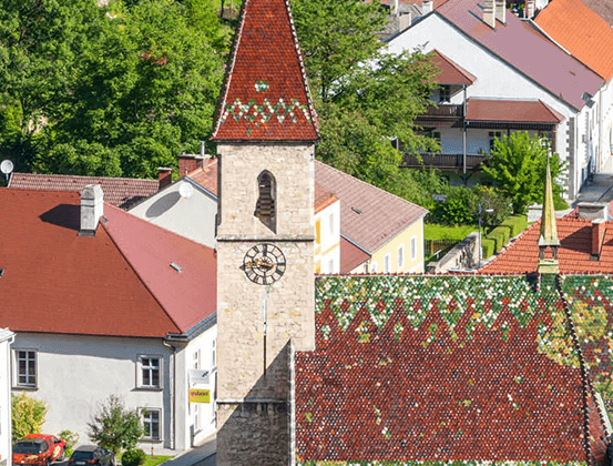 Luftaufnahme einer Stadt mit traditioneller Architektur. Rot gedeckte Dächer, ein Turm mit einer Uhr und üppige Grünflächen. Mehrere Häuser mit Schornsteinen, einige geparkte Autos und eine steile Straße.