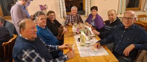 Eine Gruppe älterer Erwachsener, sowohl Männer als auch Frauen, versammelt sich um einen Tisch mit Weingläsern und einem Blumengesteck. Sie scheinen in ein Gespräch vertieft zu sein.