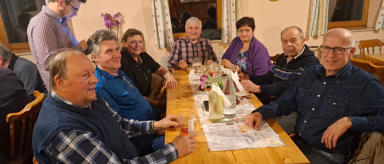 Eine Gruppe älterer Erwachsener, sowohl Männer als auch Frauen, versammelt sich um einen Tisch mit Weingläsern und einem Blumengesteck. Sie scheinen in ein Gespräch vertieft zu sein.