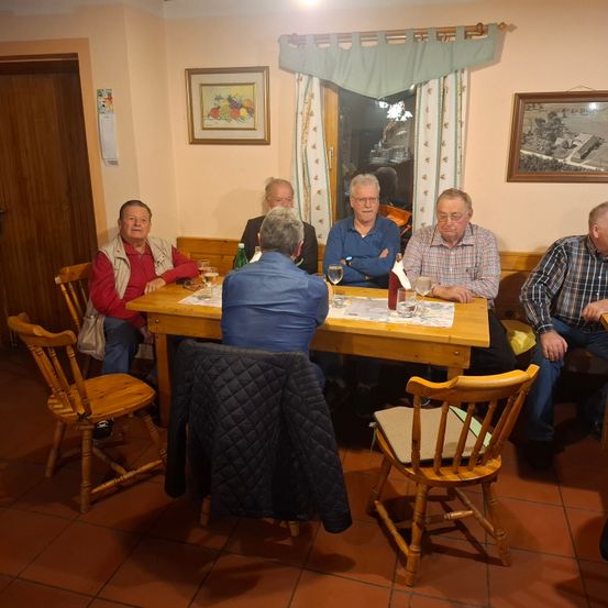 Sechs Männer sitzen um einen Holztisch in einem Raum mit beigefarbenen Wänden und trinken Wein. Ein Mantel hängt auf einem Stuhl vor ihnen. Zwei Gemälde hängen an der Wand.
