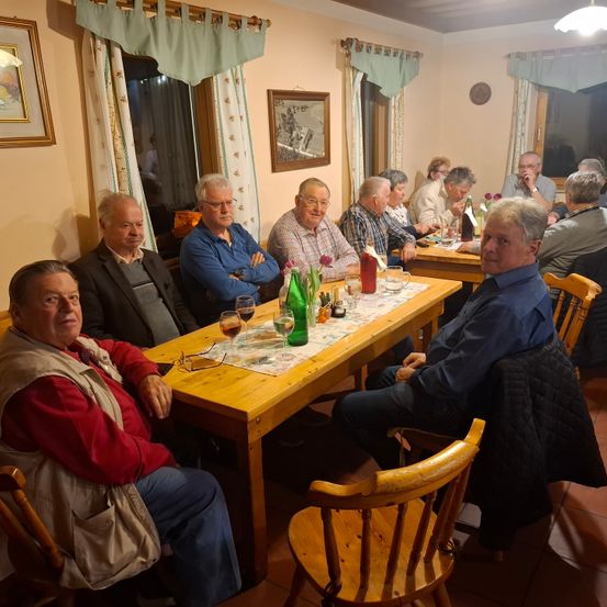 Eine Gruppe von Menschen sitzt an einem Tisch in einem Raum mit Weingläsern, Flaschen und Blumen. Einige tragen Brillen und Jacken. Zwei Fenster mit Vorhängen sind an der Wand.