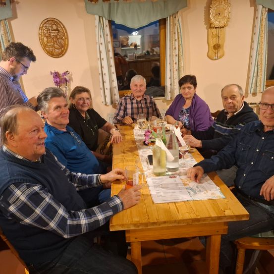 Eine Gruppe von Menschen sitzt um einen Tisch in einem Raum und lächelt. Sie scheinen eine Mahlzeit zu haben. Der Tisch hat Weingläser, Flaschen und eine Speisekarte. Die Wand hat eine Uhr und einen runden Gegenstand. Es gibt Fenster mit Vorhängen.