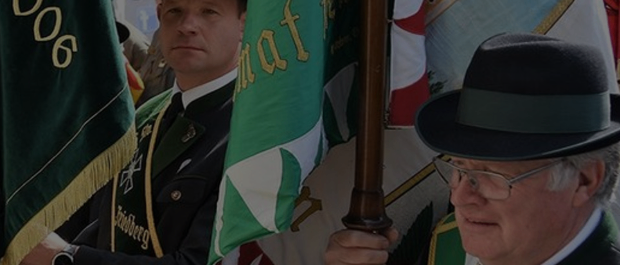 Mehrere Personen in formeller Kleidung halten Fahnen. Der Mann vorne trägt eine Brille und einen schwarzen Hut. Die Fahne, die er hält, hat grüne und weiße Abschnitte.