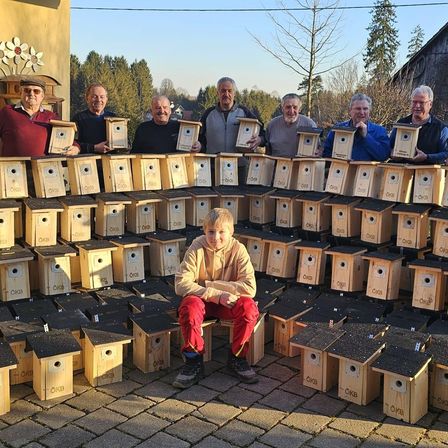 Eine Gruppe von Männern und einem Kind posiert vor einer großen Sammlung von holzverkleideten Vogelhäusern.