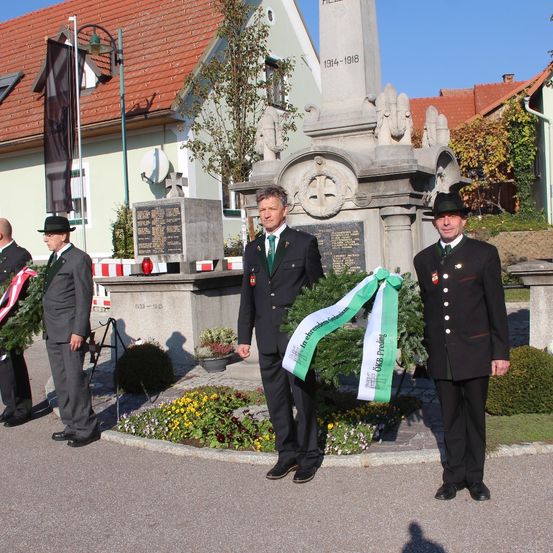 Drei Männer in Uniformen stehen neben einem Denkmal und halten Kränze. Das Denkmal ist aus Stein mit einem Kreuz und hat die Jahre 1914-1918 eingraviert.