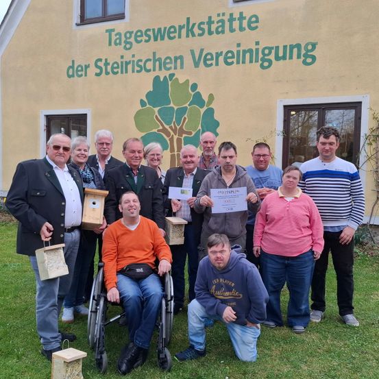 Eine Gruppe von Menschen vor einem Gebäude mit einem Baumwandbild, posiert für ein Foto mit Urkunden und Vogelhäusern.