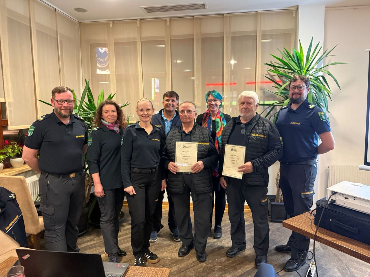 A group of people in uniforms stand in a room, posing for a photo. They hold certificates. Behind them are curtains and a plant.