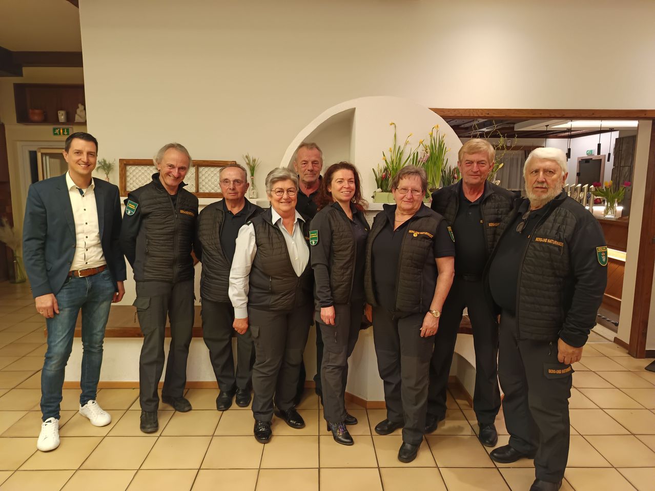 A group of adults are standing together, possibly for a photo, wearing jackets. They are smiling and appear to be posing for a picture.