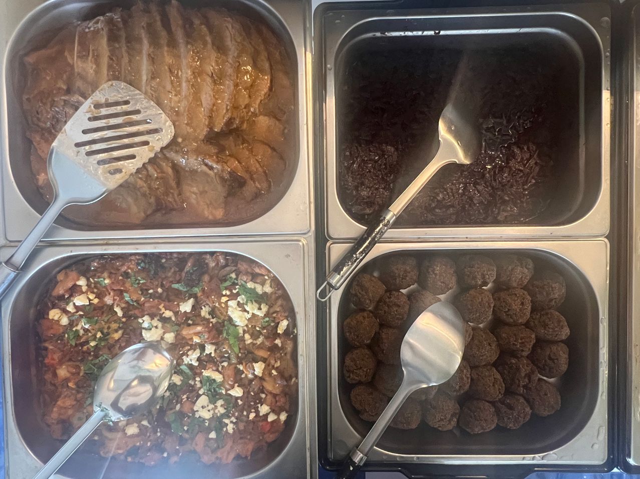 Vier Edelstahlschalen mit verschiedenen Speisen, darunter Fleischbällchen, ein Gemüsegericht und ein gebratenes Fleisch mit Sauce. Jede Schale hat einen Spatel und einen Löffel.