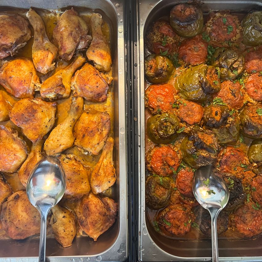 Zwei Speisentabletts: eines mit gebratenen Hähnchenschenkeln und ein anderes mit gefüllten Paprikaschoten. Beide Tabletts haben Löffel zum Servieren.