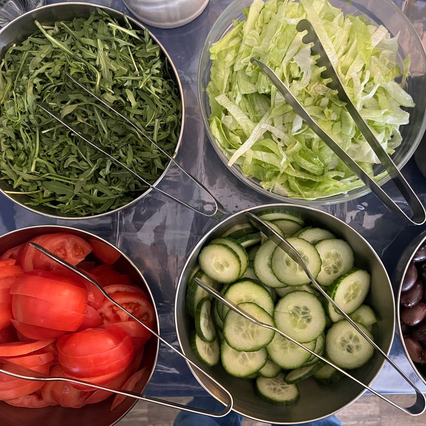 Schüsseln mit Salatzutaten wie Salat, Gurken und Tomaten. Zangen in jeder Schüssel.