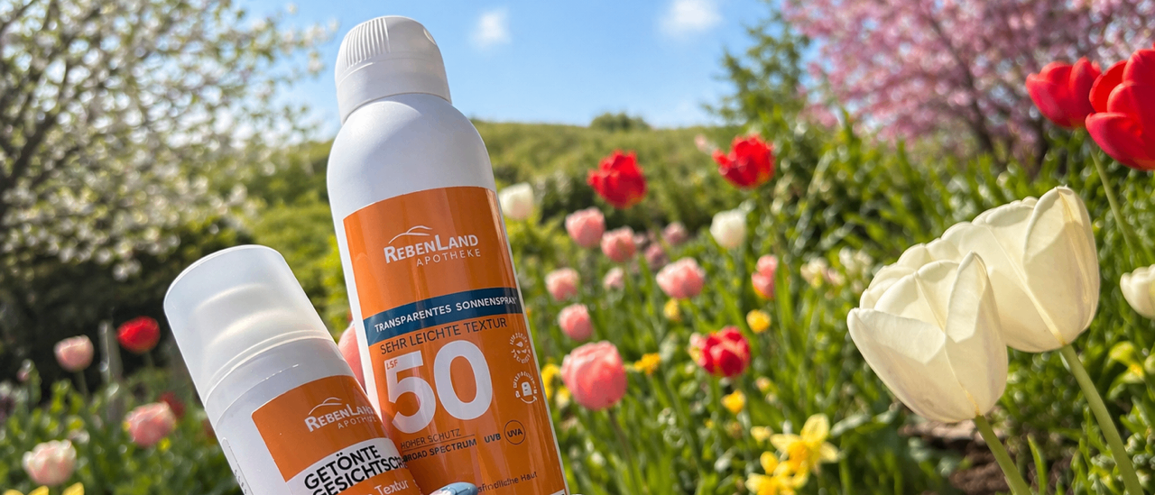 Zwei Flaschen Sonnencreme mit LSF 50 werden in einer Hand gehalten. Die Flaschen sind orange und weiß. Sie werden von einem Garten mit bunten Blumen umgeben. Der Himmel ist blau mit einigen Wolken.